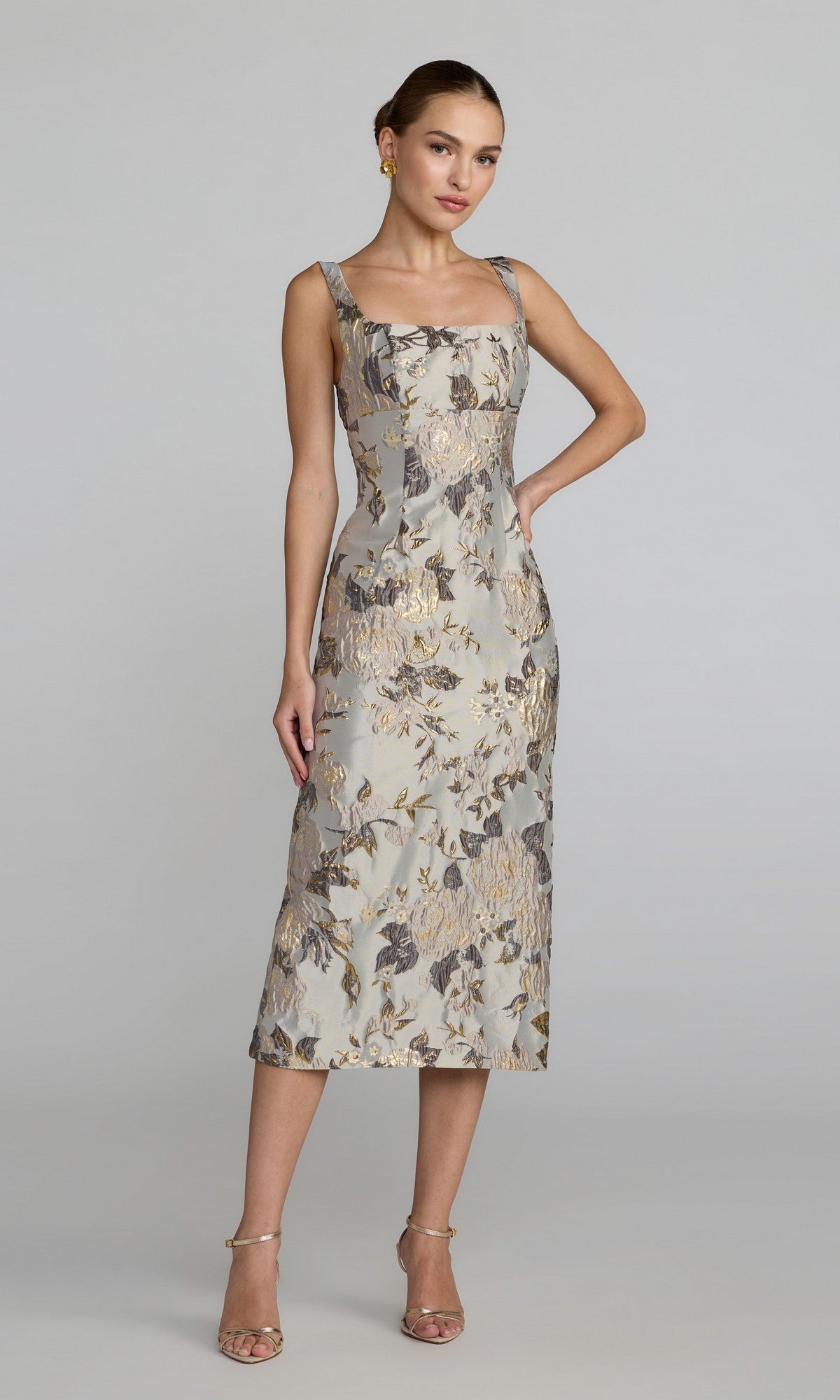 A woman stands in the Mac Duggal 69152 Floral Midi Wedding Guest Dress in Platinum Multi—a sleeveless, floral-patterned midi dress with a square neckline—styled with strappy heels and earrings against a plain background.